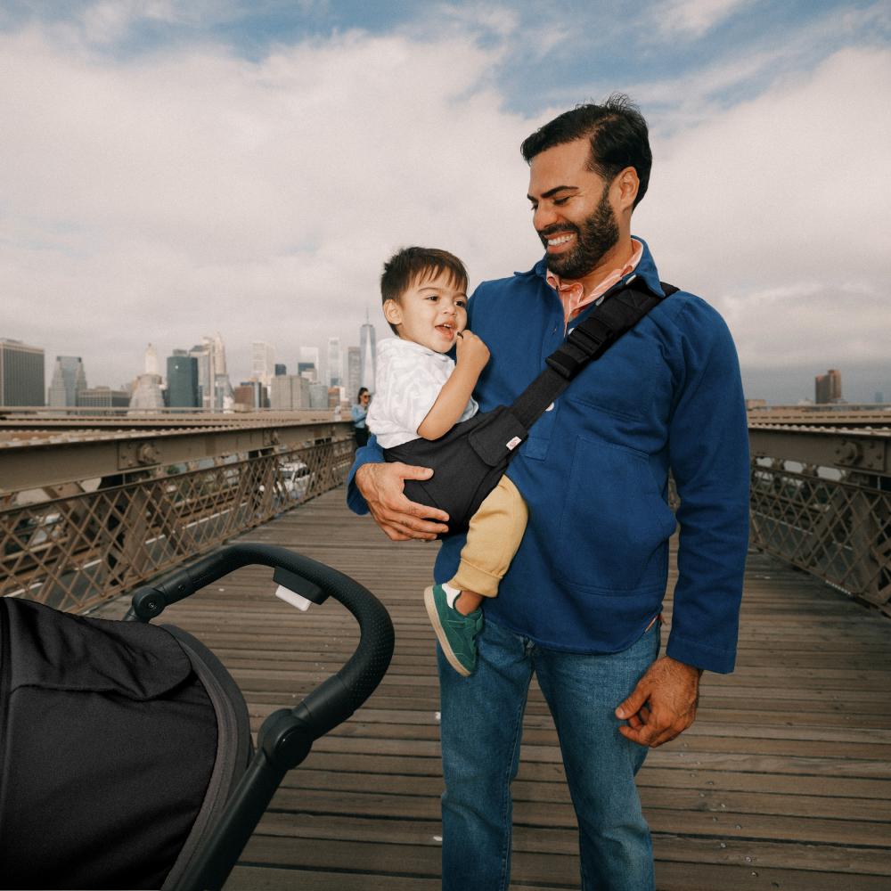 A man wearing a blue sweater and jeans carries a toddler in a black baby carrier bag across a wooden bridge, with a stroller visible in the foreground and a city skyline under a cloudy sky in the background.