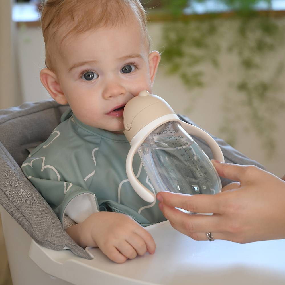 A baby wearing a sage green bib sits in a high chair while an adult hand holds a clear plastic sippy cup with a beige lid and white handle to the baby's mouth.