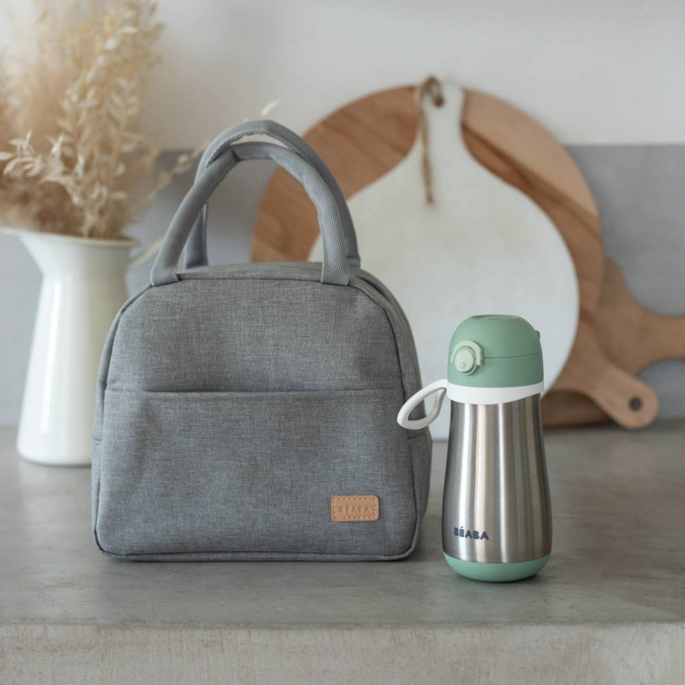 A gray fabric lunch bag with dual top handles sits on a tabletop next to a stainless steel water bottle with a mint green lid. The scene is styled with a white vase of dried grass and a wooden cutting board in the background.