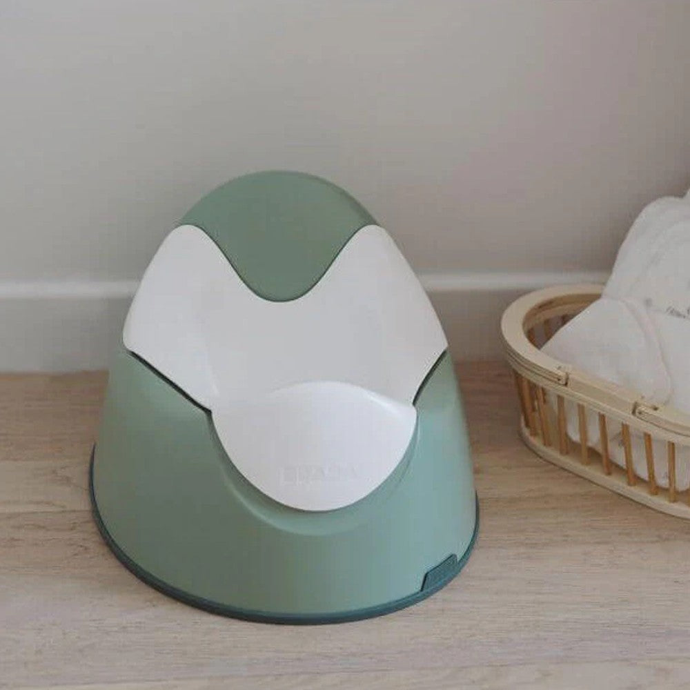 A baby potty chair in sage green and white, placed on a light wood floor next to a woven basket with a white cloth.