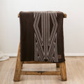 A brown baby blanket with a maori inspired pattern, draped over a wooden stool. 