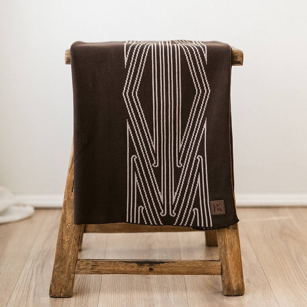 A brown baby blanket with a maori inspired pattern, draped over a wooden stool. 