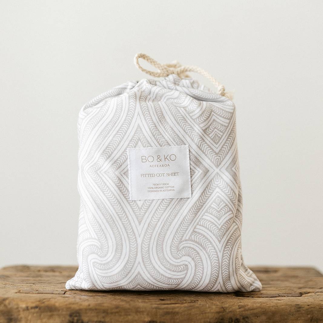 A packaged fitted cot sheet. It is white with a grey maori inspired pattern. 