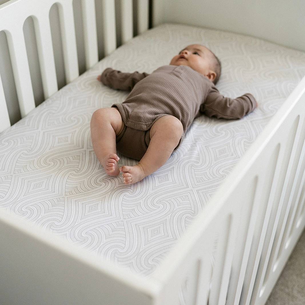 A baby lying in a cot on top of a fitted sheet that is white with a grey maori inspired pattern. 