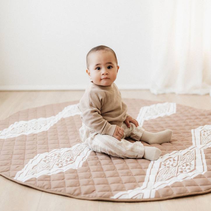 A baby sitting on a round playmat with a maori inspired design. 