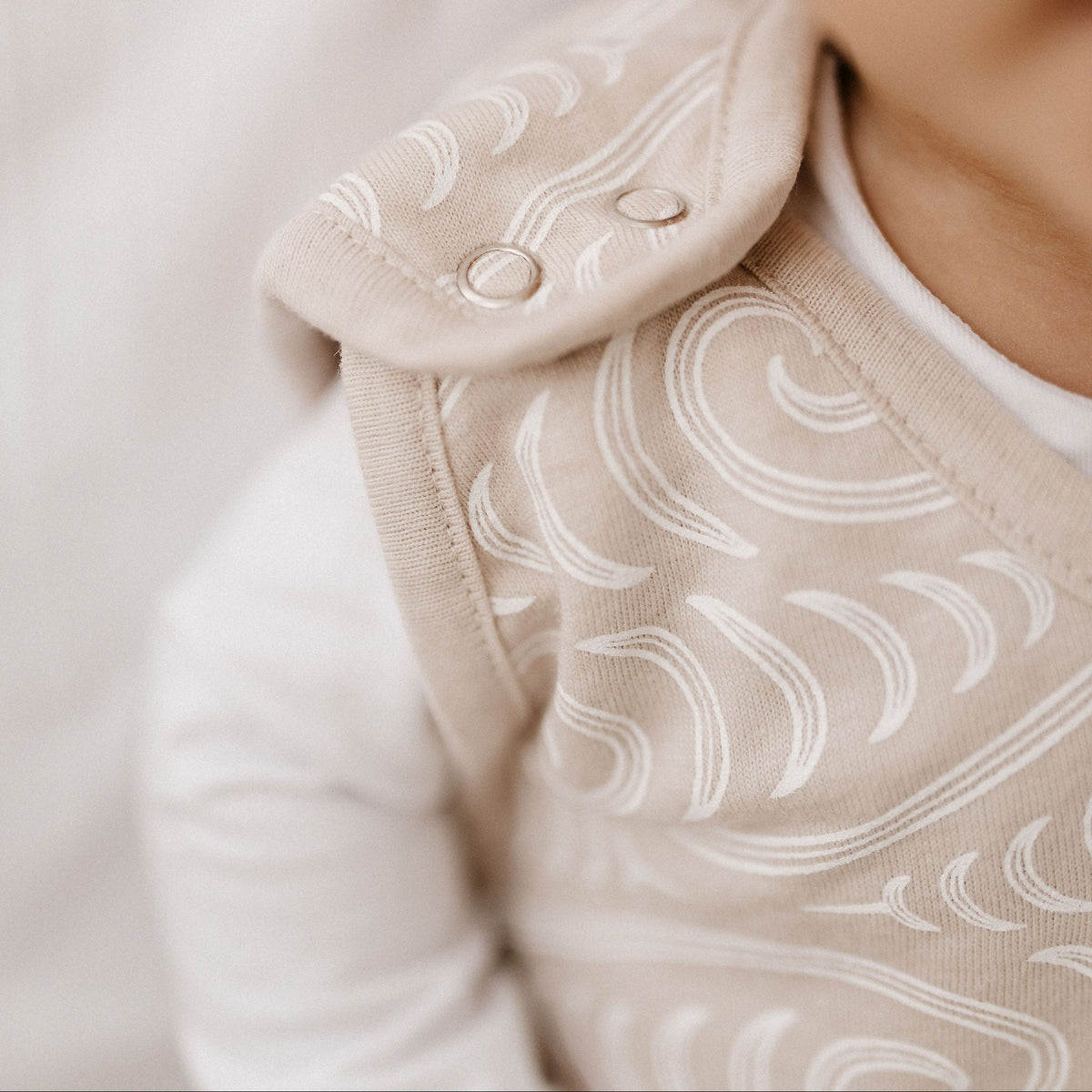 Close-up of a baby wearing a beige sleep sack with white wave patterns and snap closures, paired with a white long-sleeve shirt against a soft neutral background.