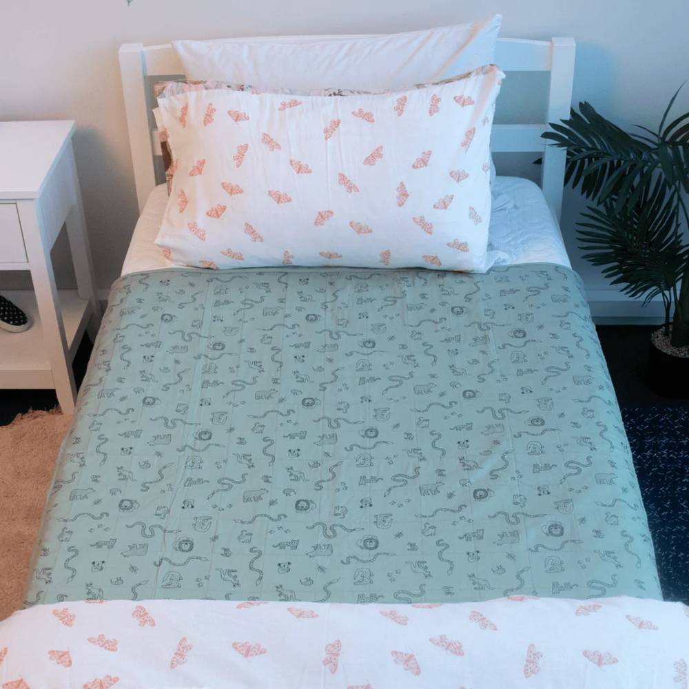 A single bed in a bedroom setting, styled with a sage green patterned brolly sheet and a white pillow featuring a pink butterfly print. A white bedside table and a potted plant are visible in the room.