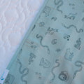 A close-up of a quilted brolly sheet with a light mint green background and a black line art animal pattern, laid next to a white quilted mattress protector