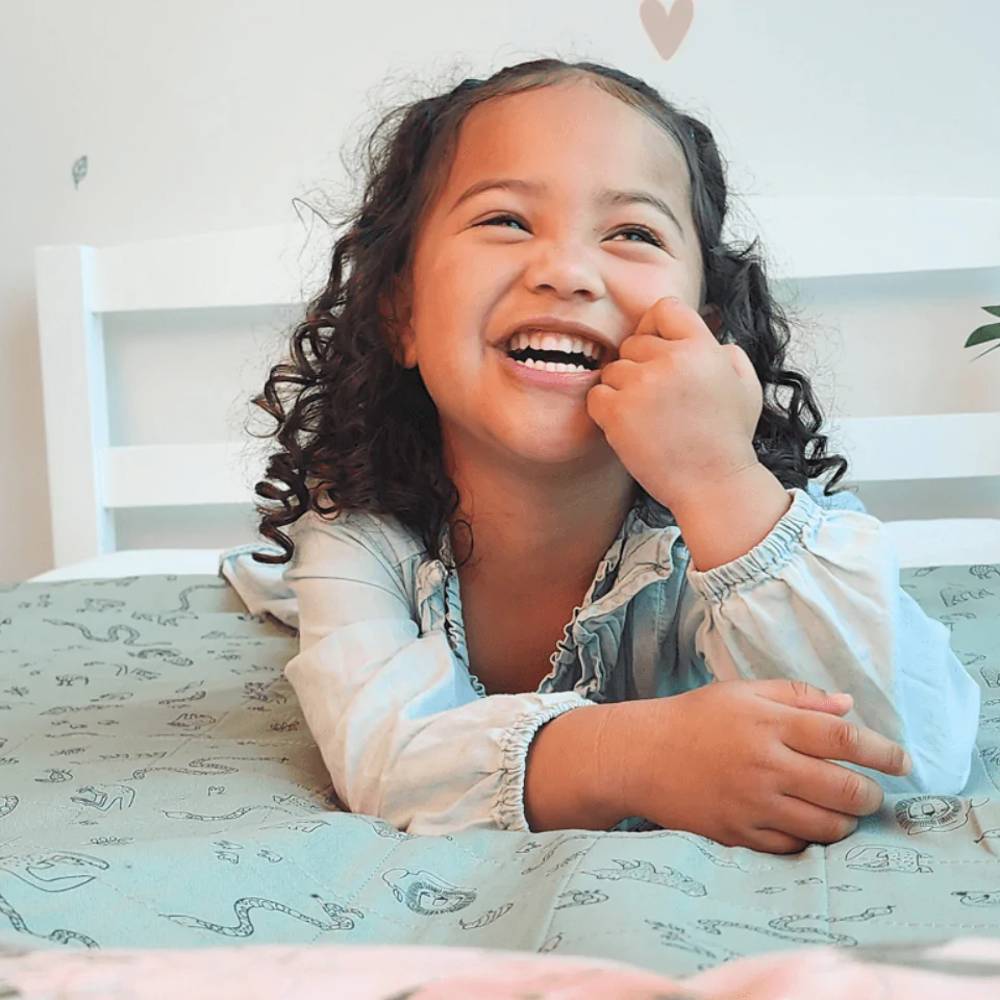 A young girl with curly dark hair smiles joyfully while lying on a bed with a light green patterned brolly sheet and white bedding.