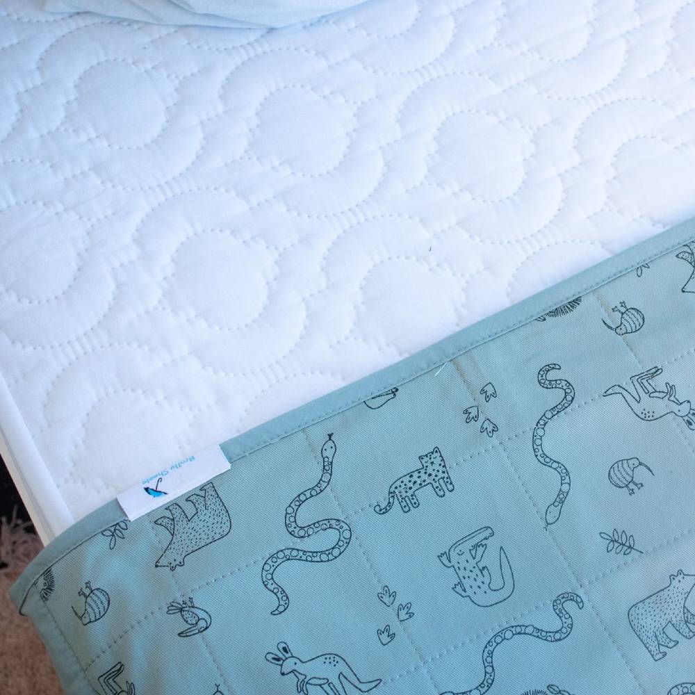 A close-up of a quilted white mattress protector bedding surface with a pale green patterned brolly sheet showing animal line drawings and a small white brand tag.