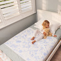 A young girl sits on a white bed wearing white pyjamas and holding a teddy bear, with a white brolly sheet featuring a light blue floral pattern underneath.