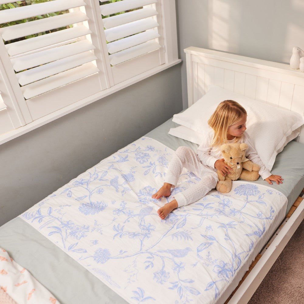 A young girl sits on a white bed wearing white pyjamas and holding a teddy bear, with a white brolly sheet featuring a light blue floral pattern underneath.