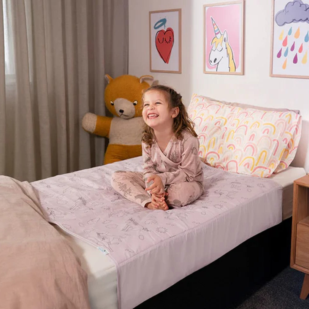 A young girl sits smiling on a bed in a child's bedroom, wearing a light pink patterned pajama set. The bed is made with a matching light pink brolly sheet with black unicorn design and a white pillow featuring a colorful rainbow design.