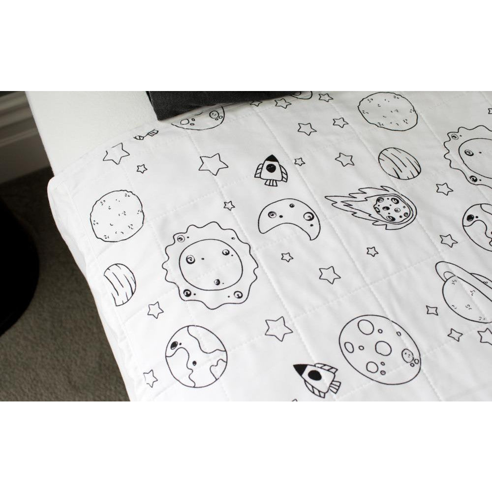 A white quilted brolly sheet featuring black line-art illustrations of space-themed motifs, including planets, moons, stars, and rockets, draped over a bed.
