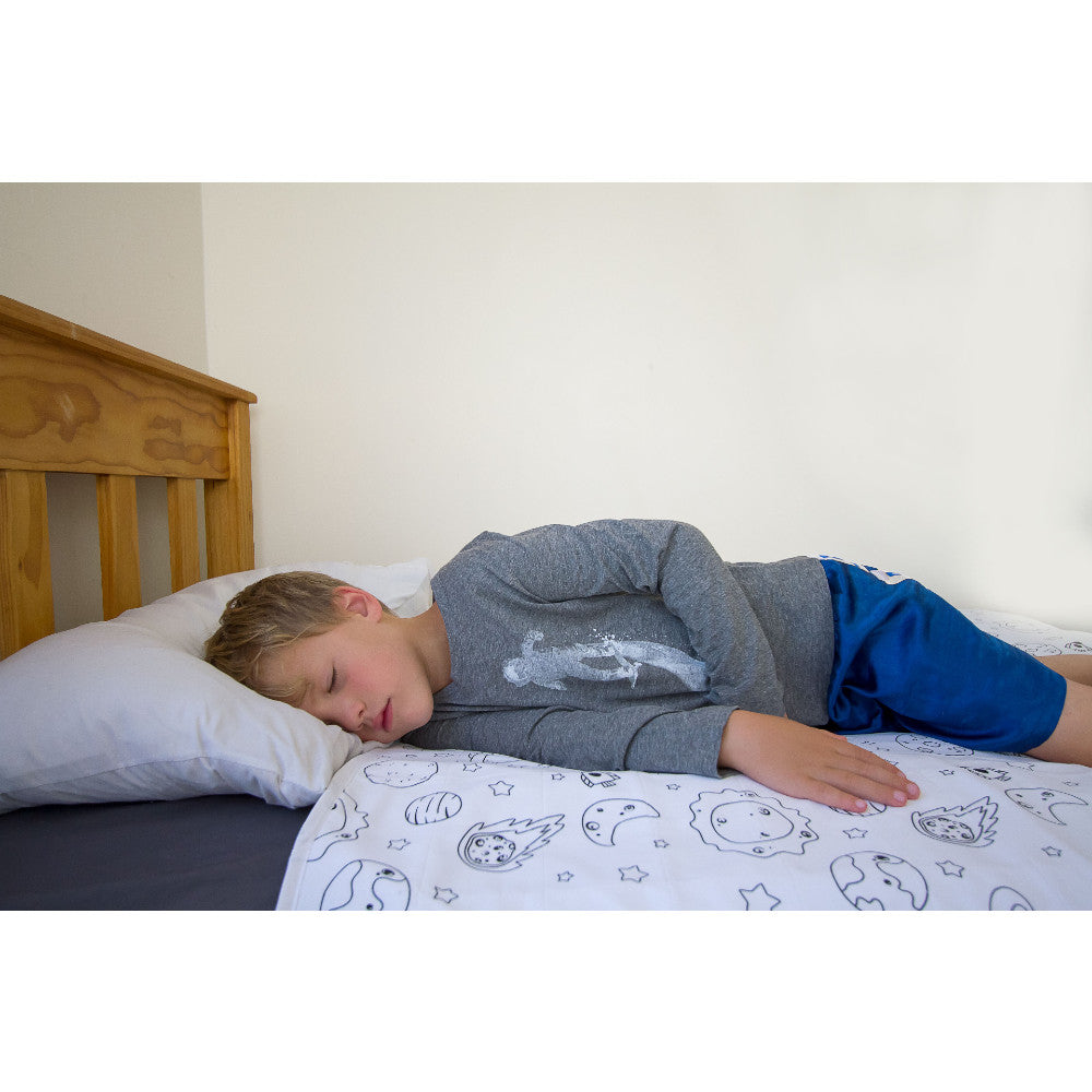 A young boy sleeping on a bed, wearing a gray long-sleeve shirt and blue shorts. The bed has a white brolly sheet with a space theme line pattern, set against a wooden headboard and a plain wall.