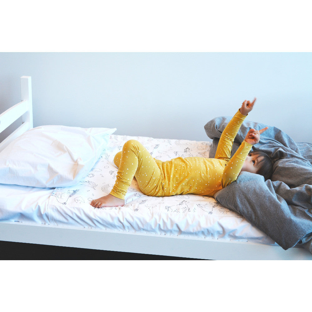 A child wearing a yellow long-sleeve pyjama set lies on their back on a white bed with patterned sheets, white pillows, and a gray blanket.