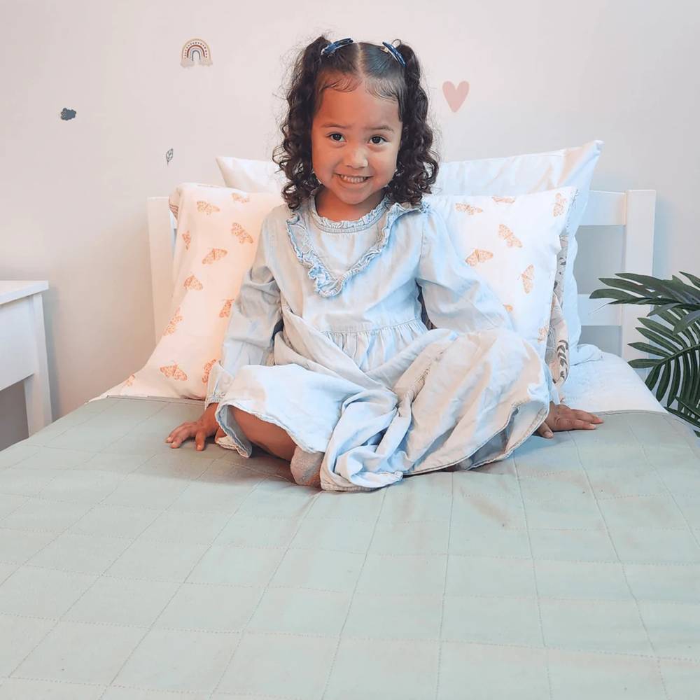 A young girl with curly dark hair sits cross-legged on a bed wearing a light blue long-sleeve dress with a ruffled collar. She is sitting on a moss green brolly sheet, surrounded by white and pale pink patterned pillows against a white headboard in a softly lit bedroom.