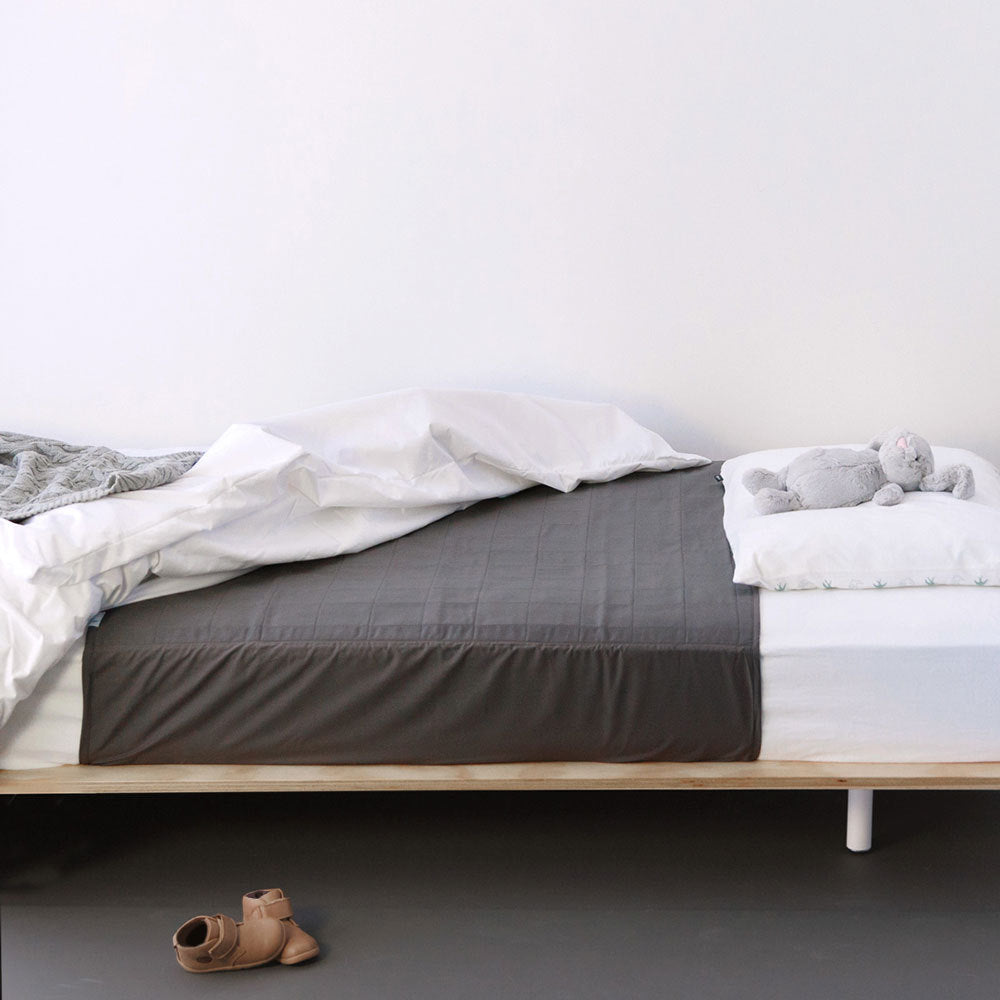 A minimalist bedroom scene showing a bed with a dark gray quilted brolly sheet and white bedding, a light wood bed frame, a small gray plush toy on the pillow, and a pair of tan slip-on shoes on the floor against a plain white wall.
