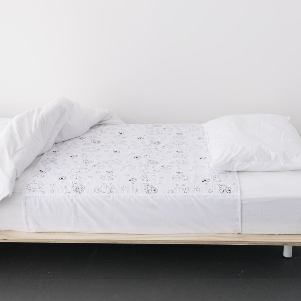 A white brolly sheet protector with black line space theme pattern is shown on a bed frame, styled with a white pillow and rumpled white duvet against a plain white wall.