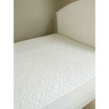 A brolly sheet white waterproof quilted mattress topper with pattern, shown on a bed with a white wooden headboard against a neutral beige wall.
