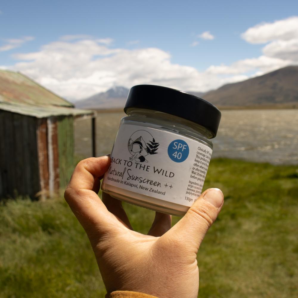 A hand holds a jar of Back to the Wild Natural Sunscreen ++ outdoors, with a grassy field, mountains, and a small wooden structure in the background under a blue sky.