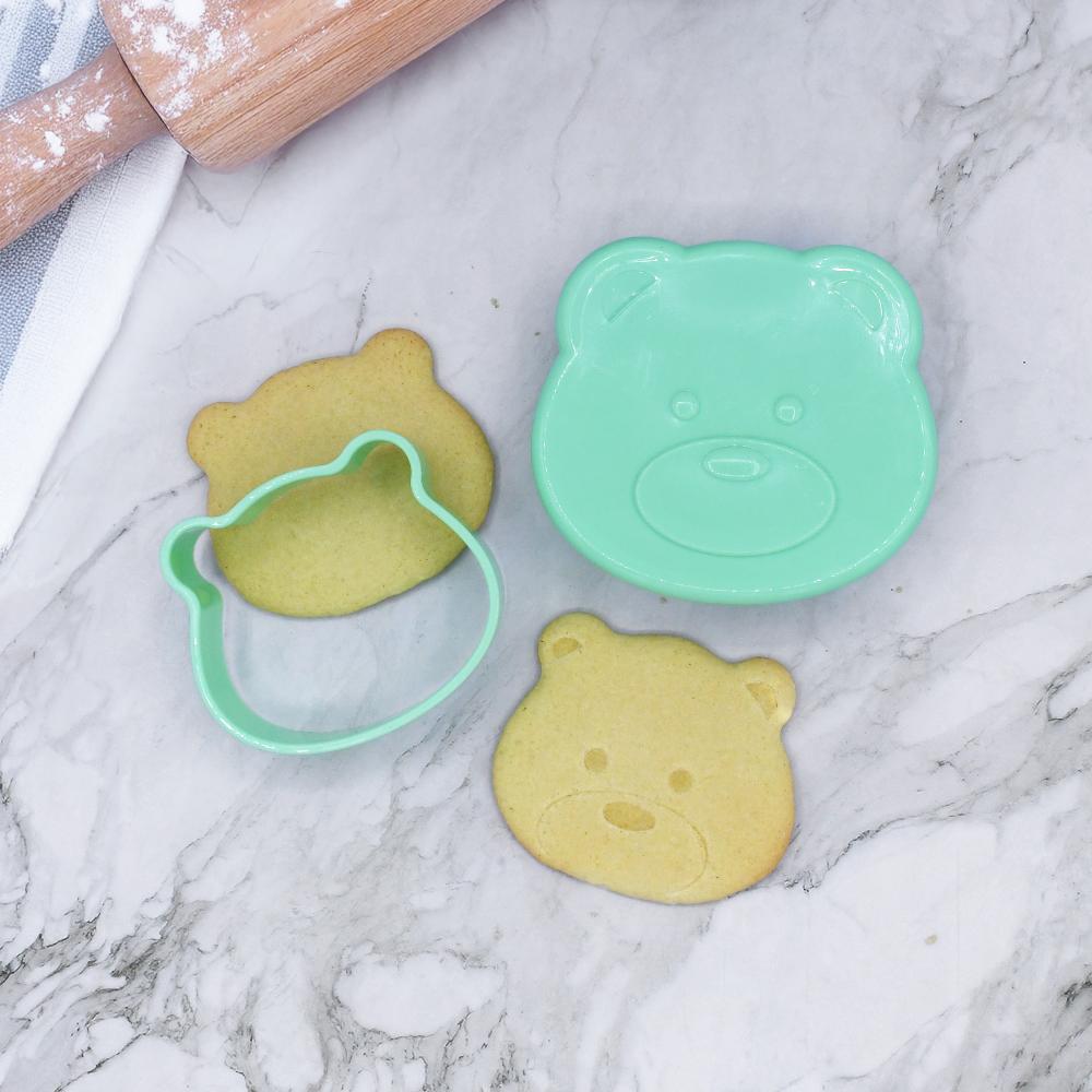 Green bear-shaped cookie cutter with two bear-shaped cookies on a marble surface.