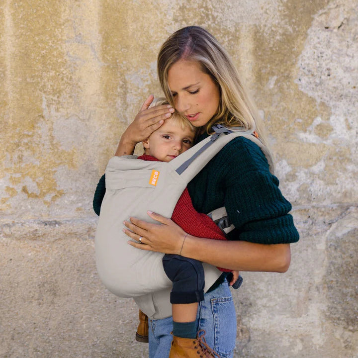 A woman wearing a dark green sweater and blue jeans carries a baby in a light gray baby carrier against a textured, weathered beige wall.