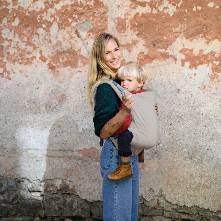 A woman wearing a dark green top and blue jeans holds a toddler in a light gray baby carrier against a weathered, peeling orange and beige wall.