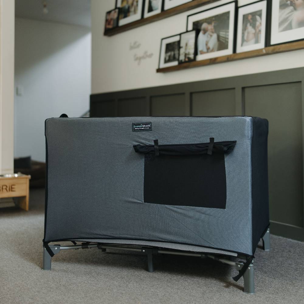 Gray Snooze Shade travel cot blackout cover in a room with framed photos on the wall.