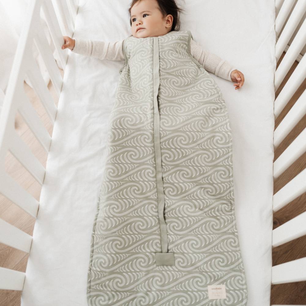 Baby lying in a crib wearing a green Maori patterned sleep bag.
