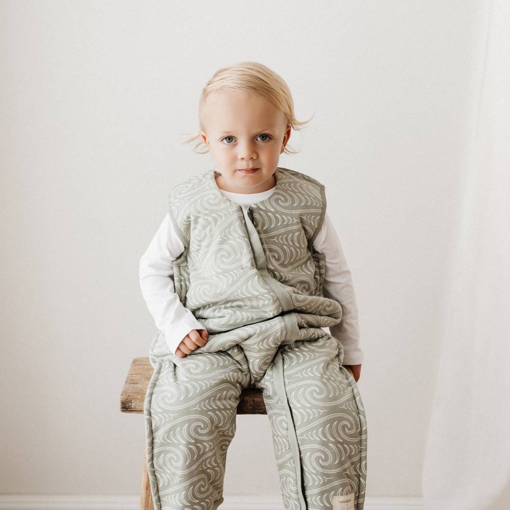 Child wearing a patterned winter sleepsuit sitting on a wooden stool against a plain background