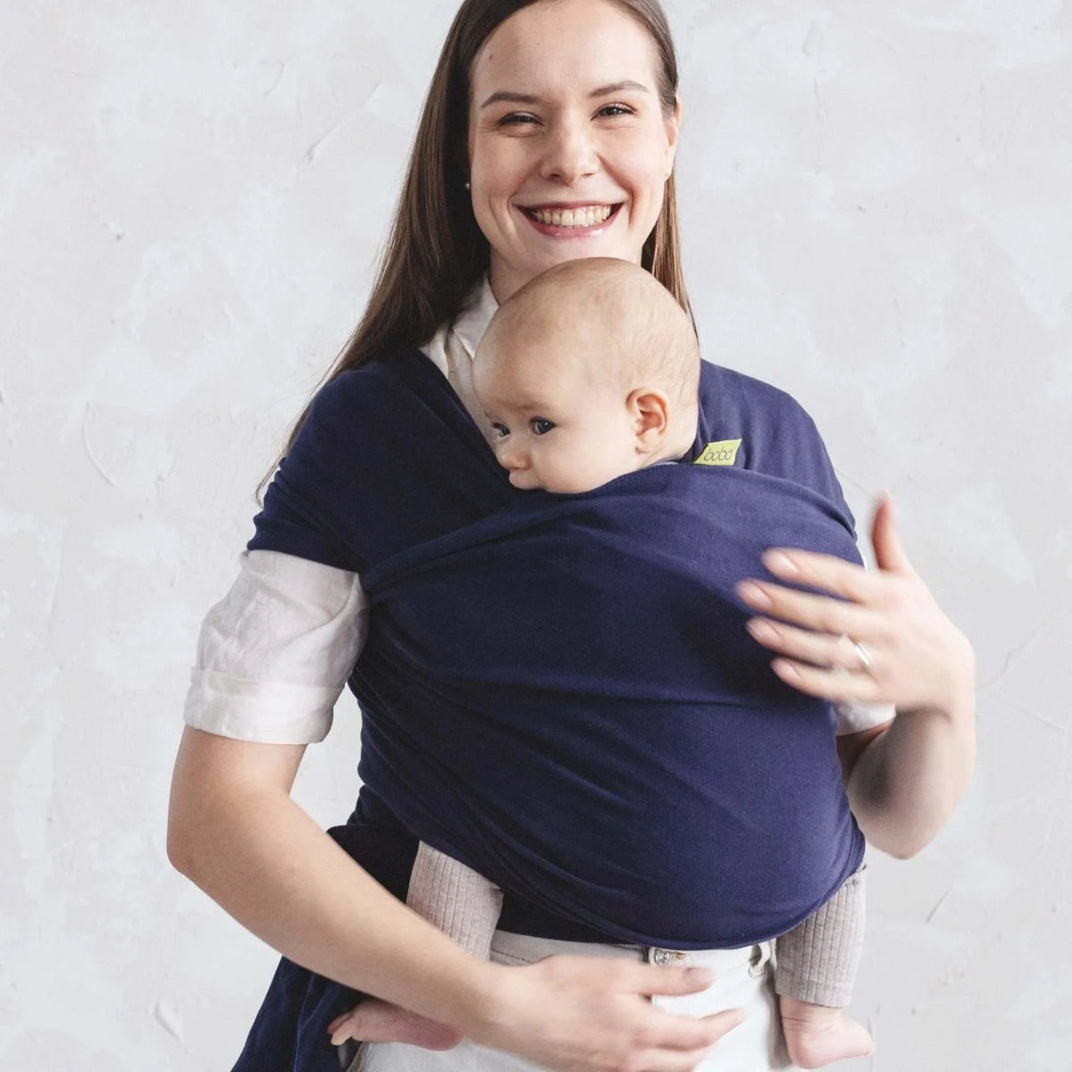 Woman smiling and holding a baby in a navy blue wrap baby carrier against a light gray background