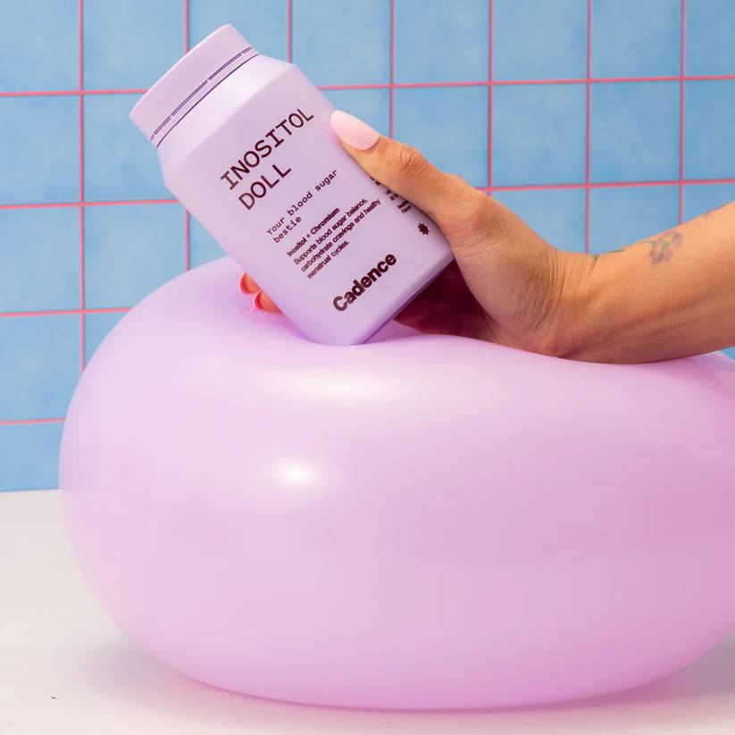 Hand holding a purple container labeled 'INOSITOL DOLL' on a purple ballooon with a grid-patterned blue background.