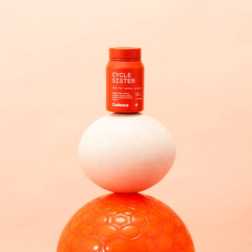 A small orange bottle labeled 'CYCLE SISTER' by Cadence, placed on top of a white sphere, with a textured orange sphere underneath, against a soft peach background.