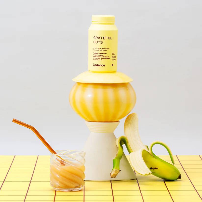 A yellow bottle labeled 'GRATEFUL GUTS' and 'Cadence' displayed on a yellow-and-white pedestal, with a peeled banana and a glass of orange drink on a yellow grid surface against a light gray background.