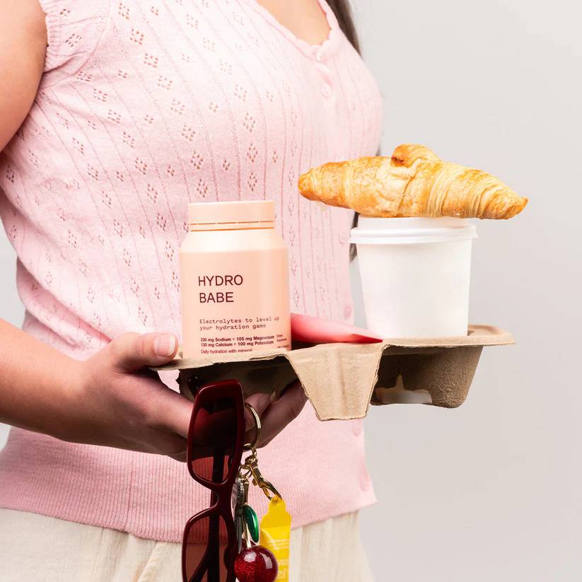 A person in a pink top holds a brown paper food tray carrying a pink 'HYDRO BABE' supplement bottle, a white coffee cup, and a croissant on top, with sunglasses and keys hanging from the handle.