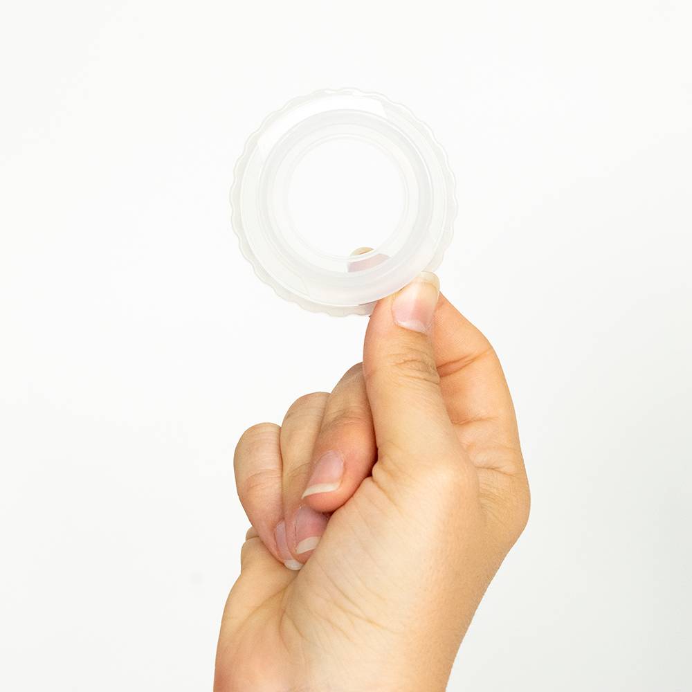 A hand holding a small, clear, circular plastic bottle adapter ring against a plain white background.