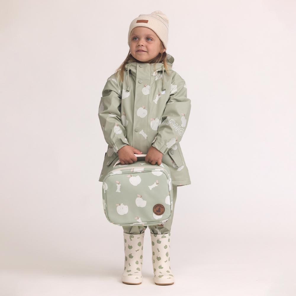 A young child stands against a plain light background wearing a sage green raincoat and matching rain pants with a white apple print, holding a matching green and white insulated lunch bag. The child also wears cream-colored rain boots and a cream knit beanie.