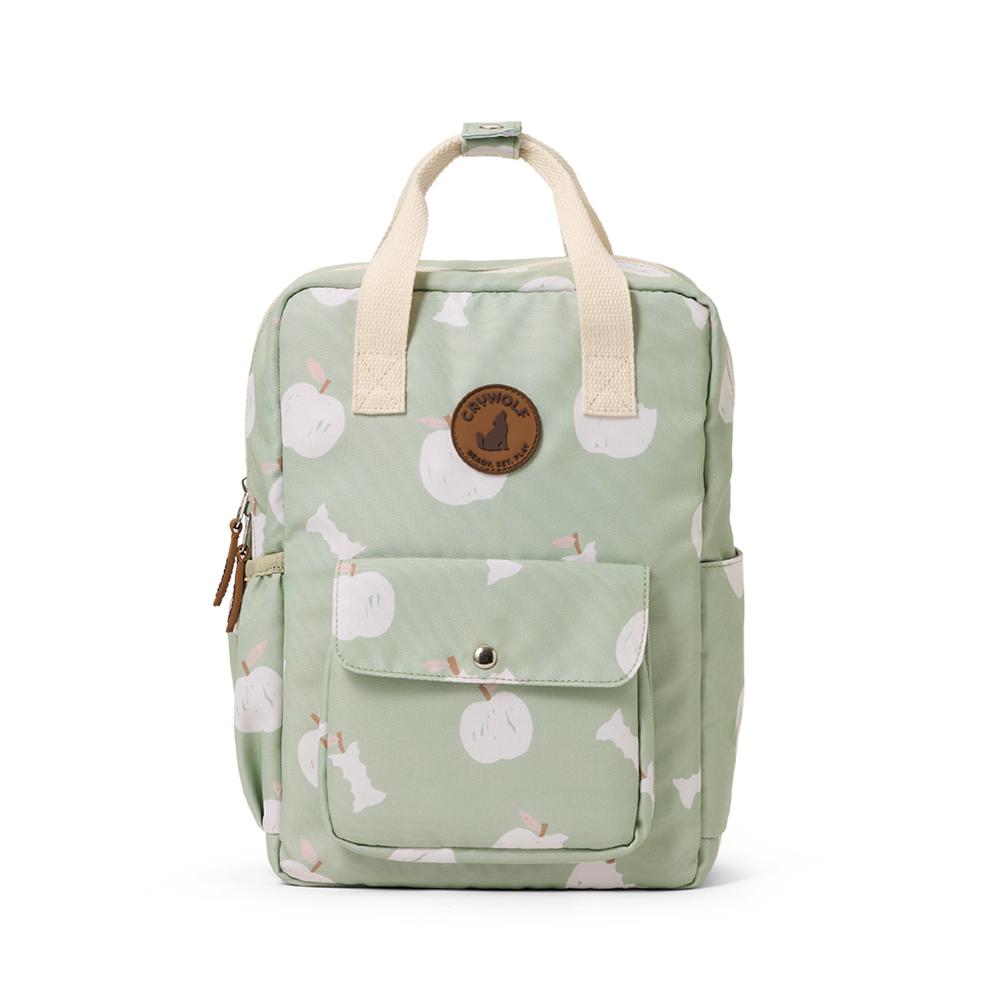 A light sage green backpack with a white apple pattern, featuring cream-colored top handles, a front pocket with a snap button, and a circular brown leather patch on the front.