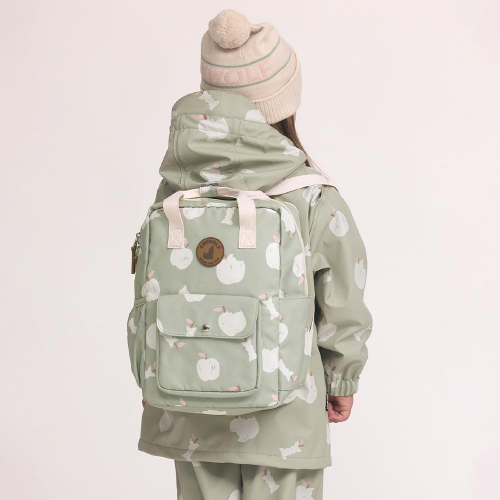A child shown from the back wearing a sage green hooded rain jacket and matching pants, and a backpack, all printed with a white apple pattern. The child is also wearing a beige knit beanie with green stripes and a pom-pom.
