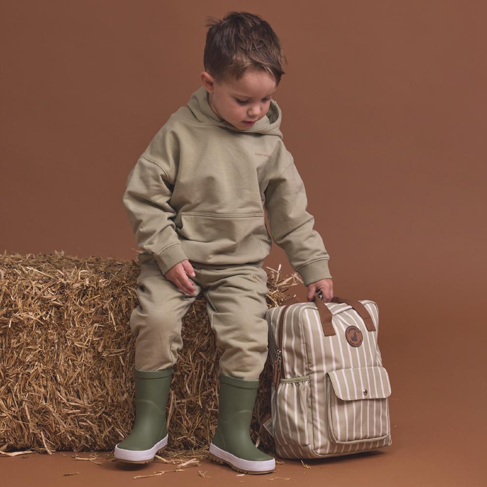 A young boy wearing a matching khaki hoodie and sweatpants set with green rain boots sits on a hay bale next to a beige and white striped backpack against a solid brown studio background.