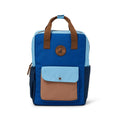 A color-blocked backpack in blue, light blue, and brown, featuring dual top handles, a front pocket with a snap closure, and a circular brown logo patch on the upper front.