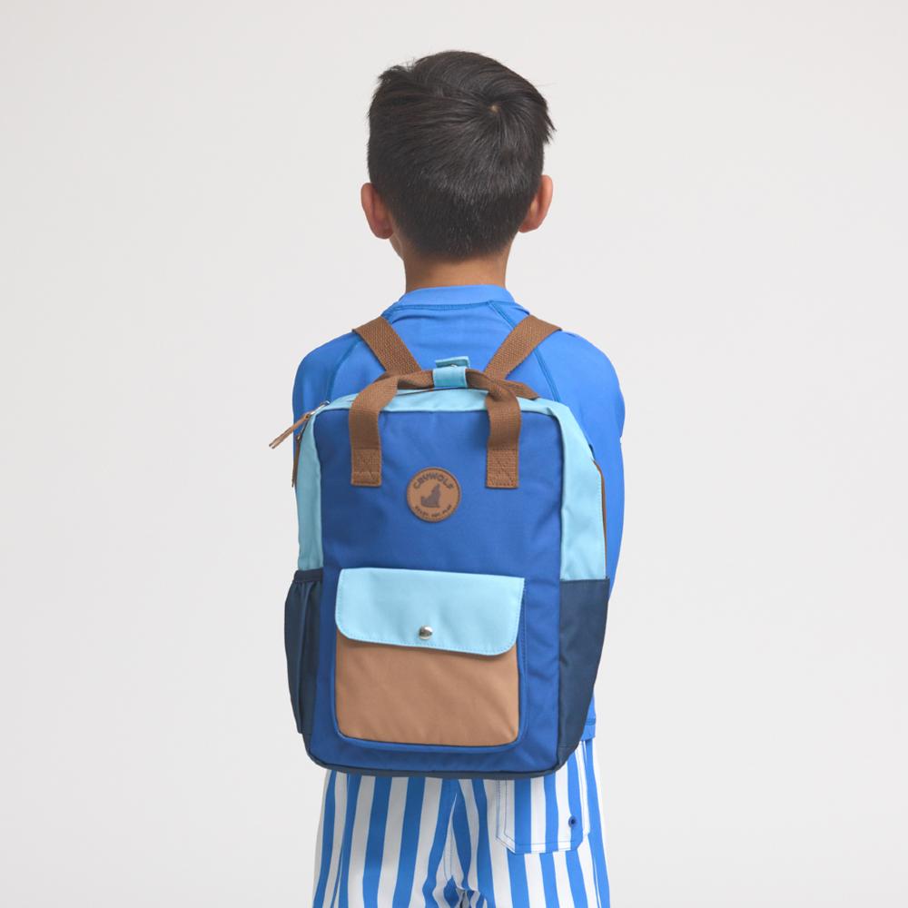 A child viewed from behind wearing a blue backpack with brown straps and a light blue front pocket, standing against a plain light gray background.