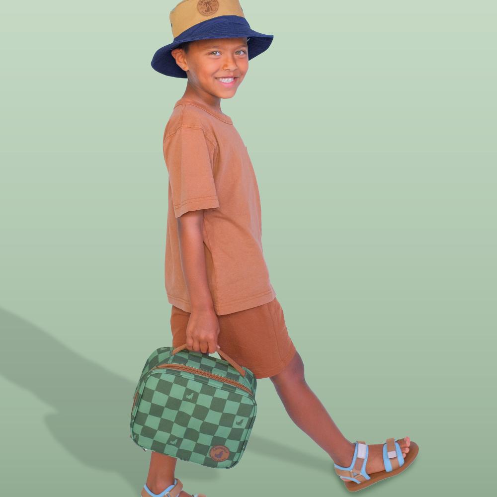 A child wearing a tan and navy bucket hat, brown t-shirt, and brown shorts walks while carrying a green and brown checkered lunch bag against a solid light green background.
