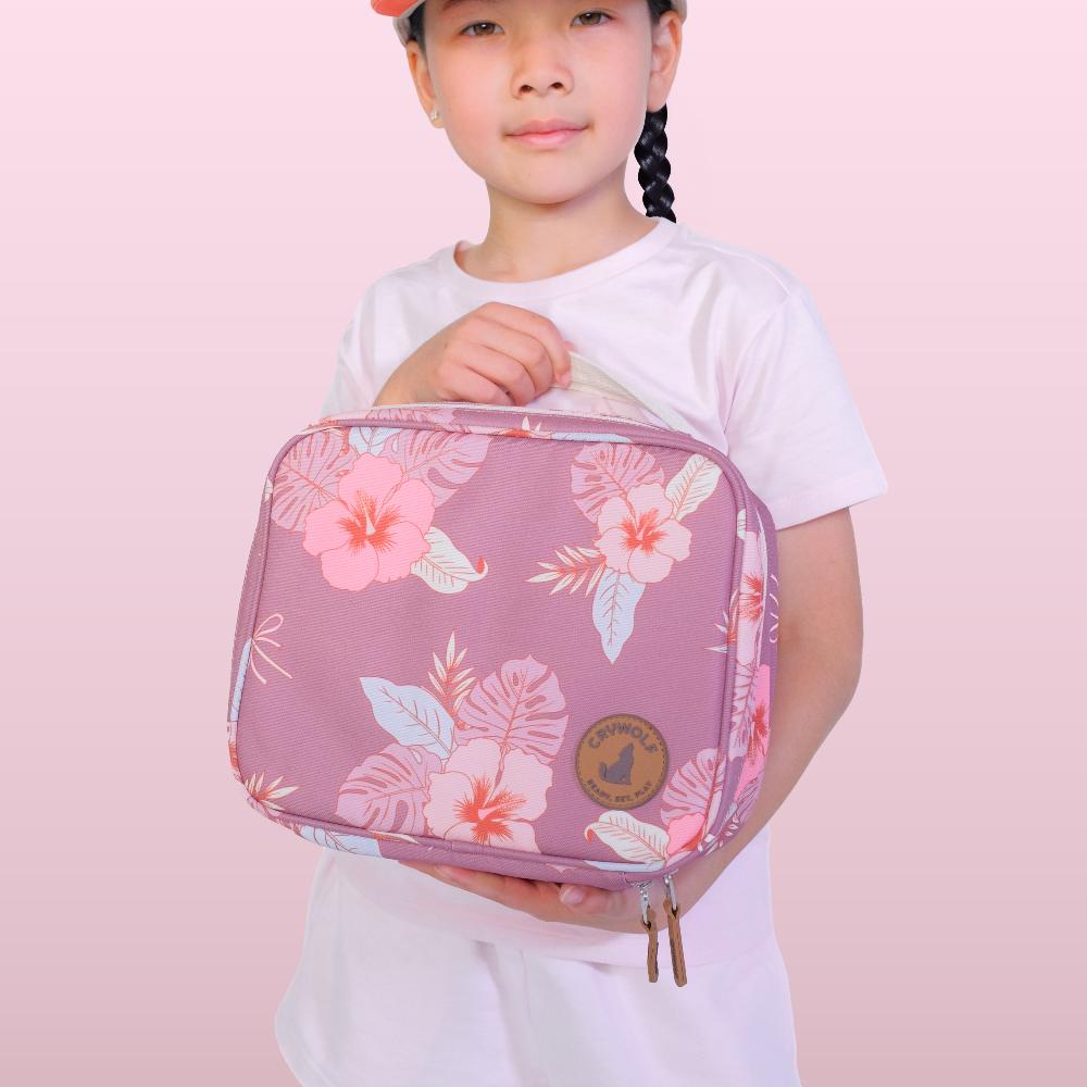 A young girl with braided hair wearing a white t-shirt and a pink cap holds a pink floral zippered lunch bag against a solid light pink background.