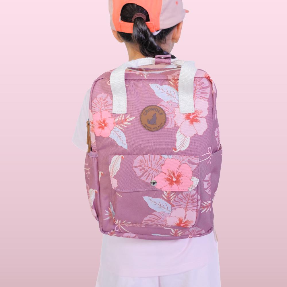 A child viewed from behind wearing a pink floral backpack with white shoulder straps and a round brown logo patch on the back, against a soft pink gradient background.