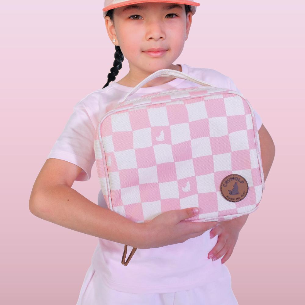 A young girl holding a pink and white checkered lunch bag, set against a solid pink background.