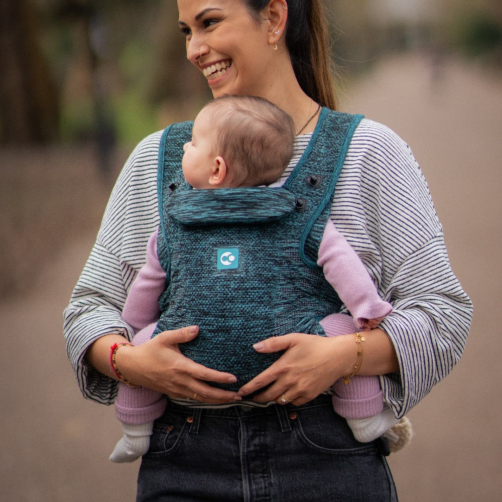 A woman wearing a dark teal woven baby carrier with a baby inside, standing outdoors on a blurred path.