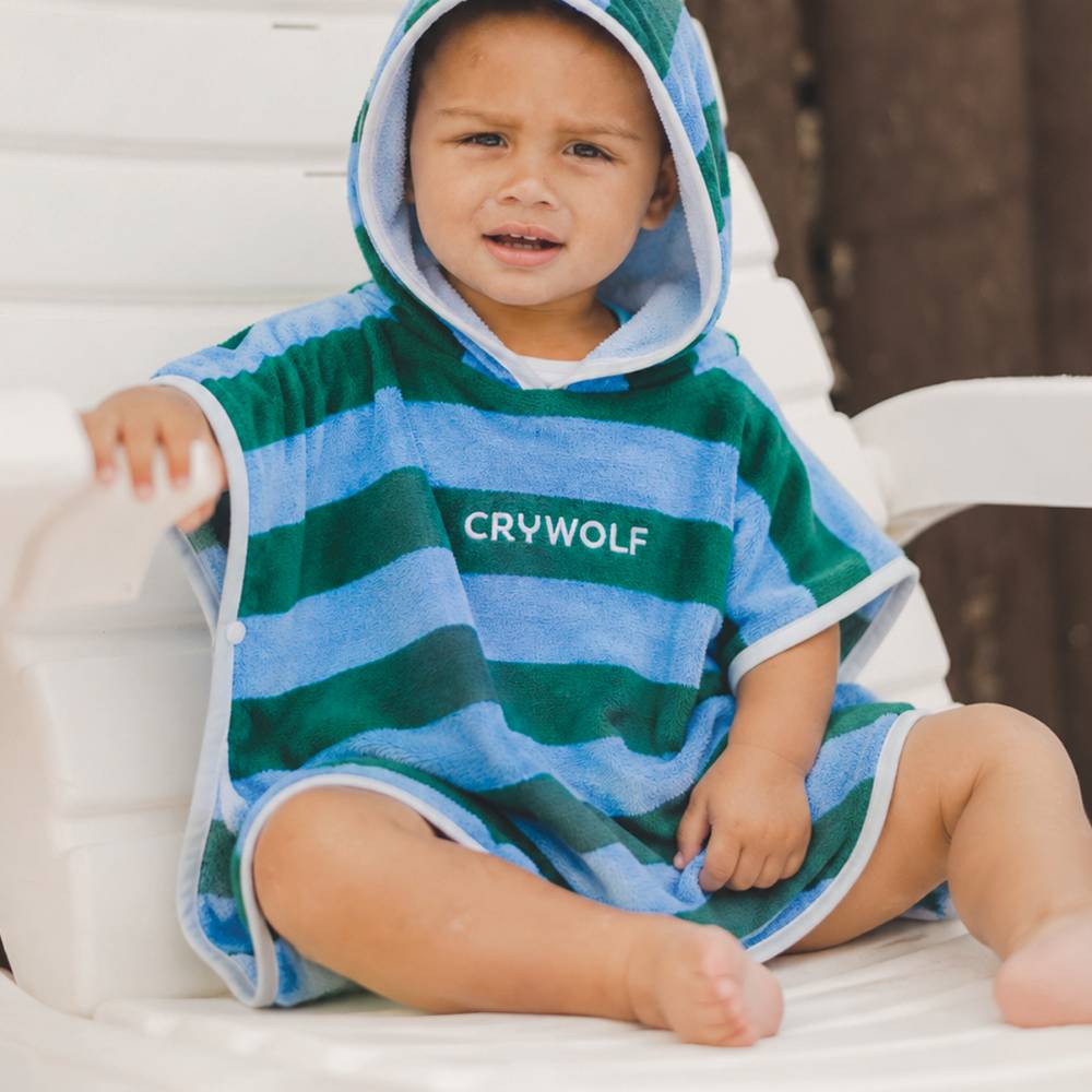 A young child wearing a green and blue striped hooded towel with the brand name 'CRYWOLF' printed on the chest, sitting on a white outdoor lounge chair.