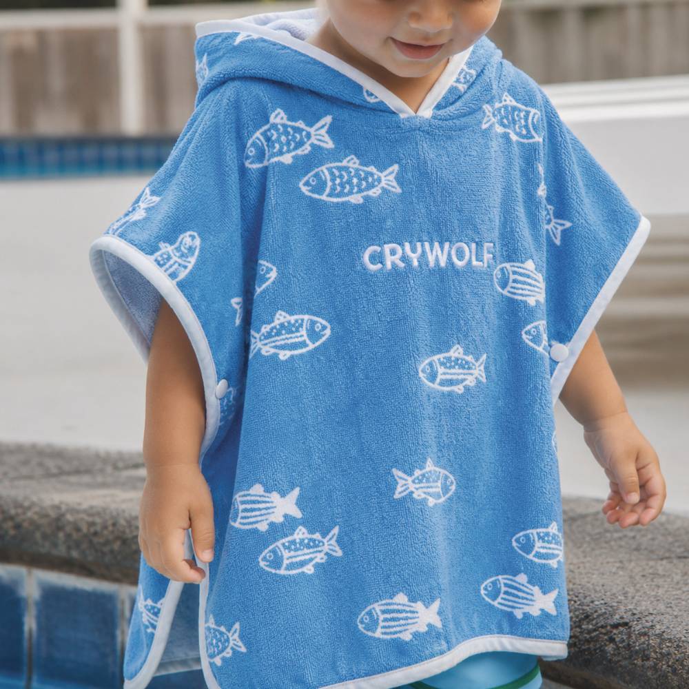A child wearing a blue hooded towel poncho with a white fish pattern and the brand name 'CRYWOLF' printed on the front, standing near a pool.